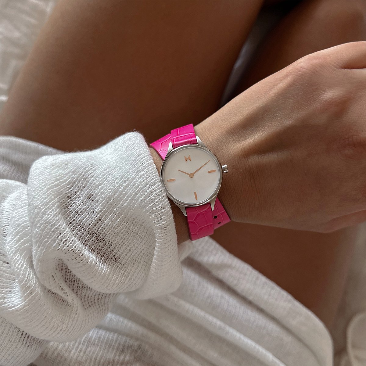 Minimalist silver-tone watch with a white dial and bright pink textured strap, worn with a white sweater