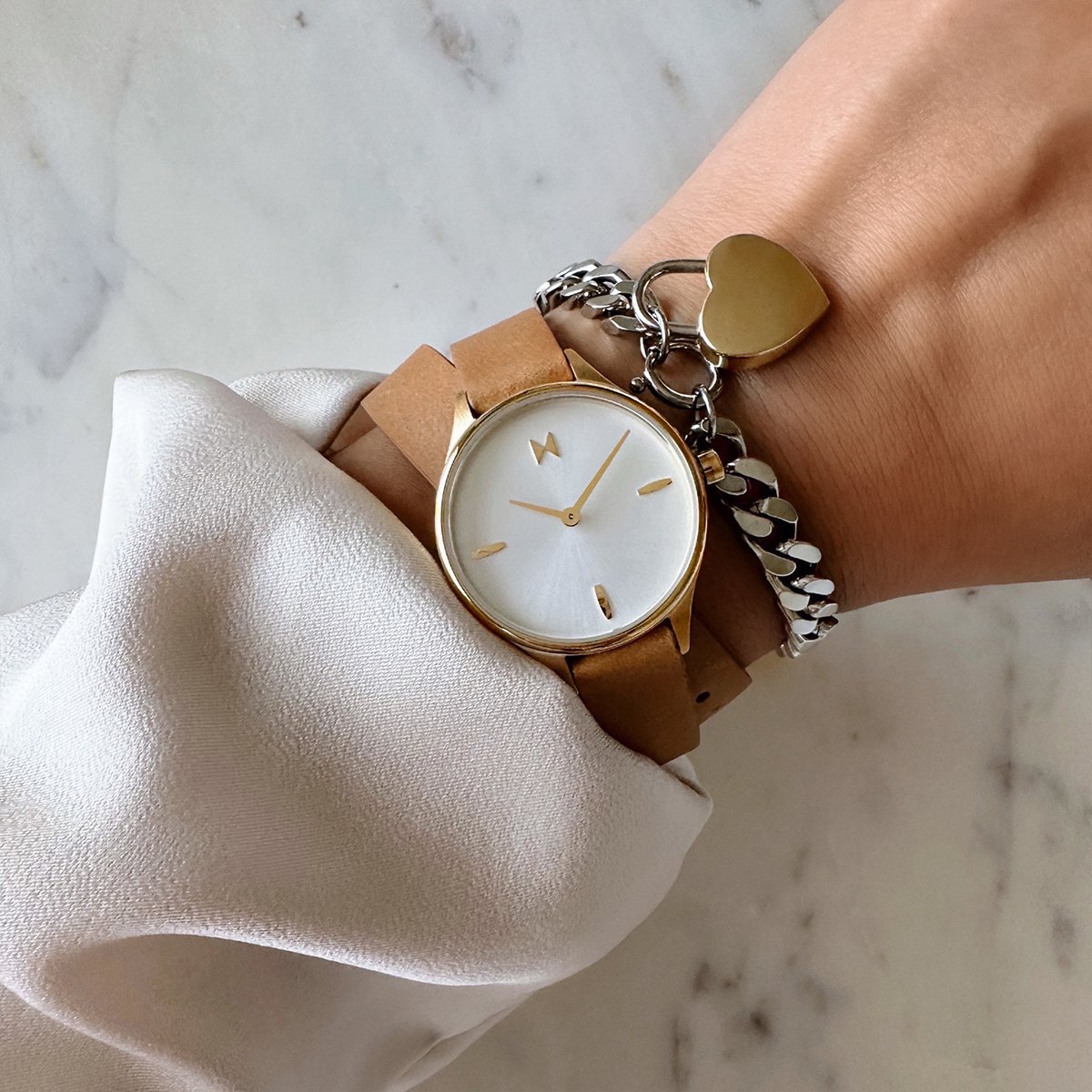 Gold-tone watch with a white dial and tan leather strap, layered with a silver chain bracelet and gold heart charm.