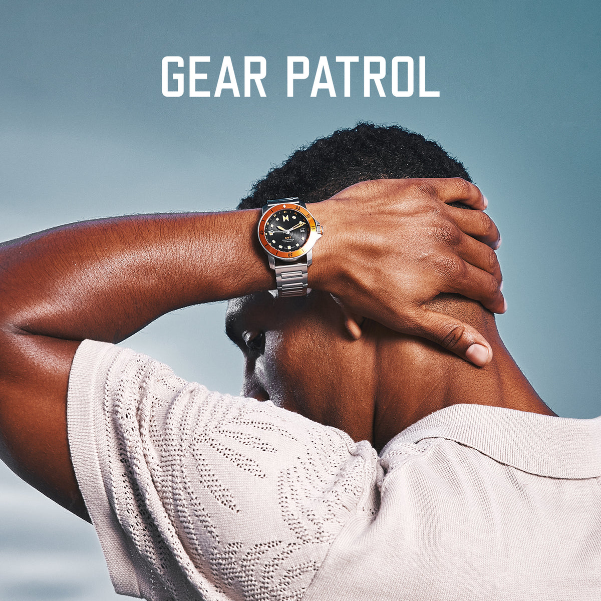 Man with his hand behind his head wearing the stainless steel Cali Diver bracelet watch with black dial and orange minute track. Featuring Gear Patrol logo at the top of the image.