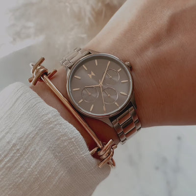 Close-up video of a gray chronograph watch with a metal bracelet worn alongside rose-gold bangles as light moves across the dial.