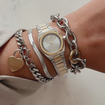 Two-tone round watch shown on a wrist layered with silver bracelets and a gold heart charm, with shifting reflections across the dial.